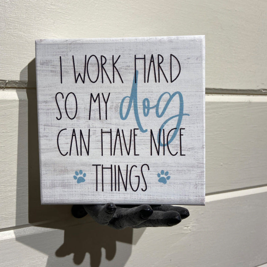 The "Have Nice Things" handcrafted decorative sign by SincereSurroundings showcases a humorous message, "I work hard so my dog can have nice things," with the word "dog" and paw prints highlighted in blue. Ideal for shelf display, it is elegantly held by a black statue of a hand against a light-colored wall in Rock Rapids, IA.