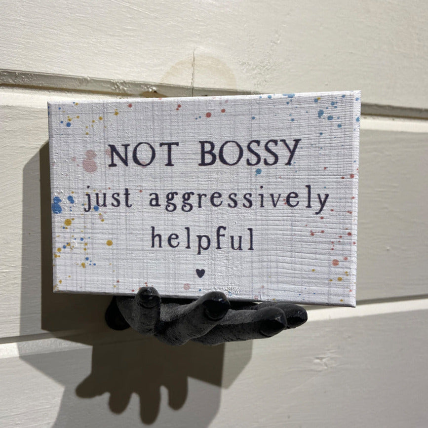 A small, handcrafted sign named "Not Bossy" by SincereSurroundings features a speckled pattern and the words "NOT BOSSY just aggressively helpful." It is elegantly displayed on a wall-mounted decorative hand for easy showcasing.