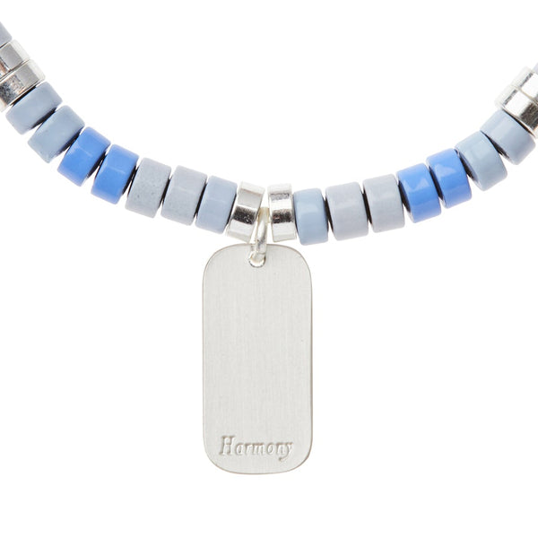 The Intention Charm Howlite Silver necklace by Scout Jewelry showcases alternating light blue, dark blue, and silver cylindrical beads. It is highlighted by a sterling silver dipped charm, with a central rectangular pendant engraved with "Harmony" to radiate positive intention.