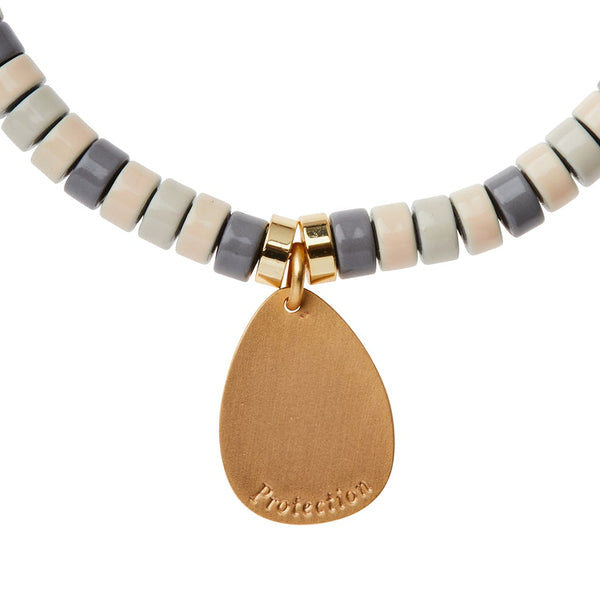 A close-up of the Intention Charm Amethyst Gold necklace from Scout Jewelry showcases its alternating gray, beige, and white semi-precious stones. The piece is highlighted by a 14k gold-dipped drop-shaped pendant engraved with the word "Protection," elegantly attached with a small gold ring to channel positive intention.