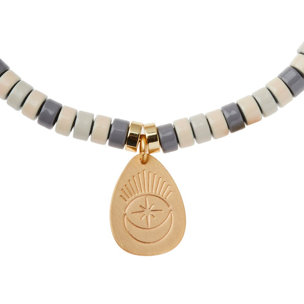 A close-up view of the Scout Jewelry Intention Charm Amethyst Gold necklace, featuring light grey, dark grey, and beige cylindrical beads. The necklace is adorned with a 14k gold-dipped charm engraved with a crescent moon under a starburst design, crafted with positive intention and semi-precious stones.