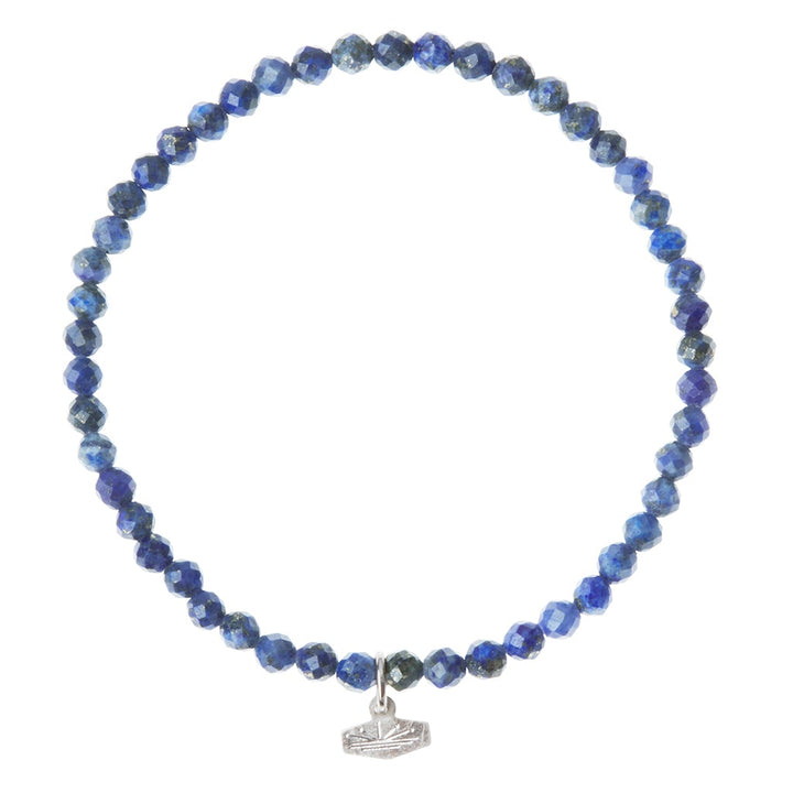Introducing the Mini Bracelet Lapis/Silver by Scout Jewelry: a stunning beaded bracelet crafted from small, semi-precious faceted lapis beads in varying shades of blue. This exquisite piece features a delicate geometric silver charm that elegantly hangs from the tightly strung, circular formation of the beads.