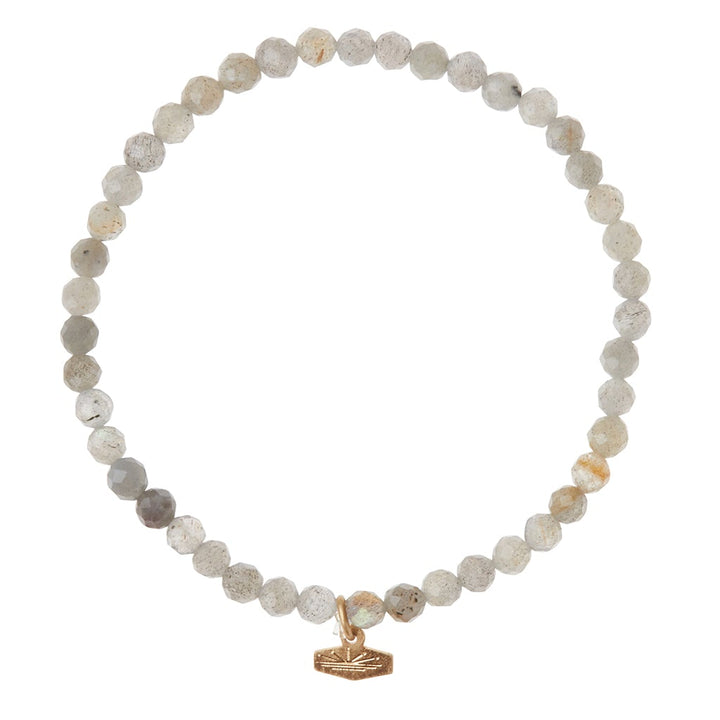 The Mini Bracelet Labradorite/Silver from Scout Jewelry is a delicate beaded bracelet crafted from translucent, light gray, faceted labradorite semi-precious gemstone beads. It features a small, silver-colored charm shaped like a rectangular tag dangling from the bracelet. The uniform-sized beads are strung on a stretch cord, forming a perfect circle.