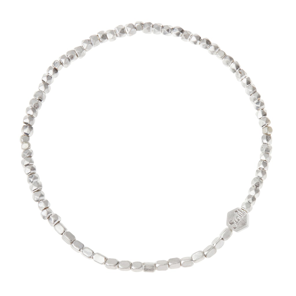 A close-up image of the Scout Jewelry Mini Metal Bracelet Silver, featuring small, irregularly shaped silver beads strung on a stretch cord. The circular bracelet includes a small hexagonal charm with text on it and showcases mixed metals for subtle accents against a plain white background.