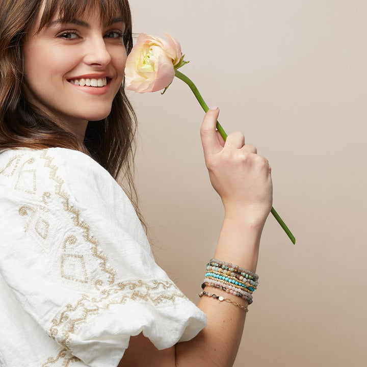 A smiling woman holds a pink flower near her face. She wears a white blouse with embroidered patterns and multiple colorful beaded bracelets, including the Chain Bracelet Aqua Terra/Silver by Scout Jewelry, adorned with semi-precious gemstones on her wrist. The background is a plain, soft beige color.