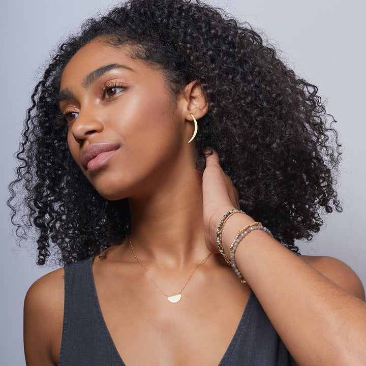 A person with curly hair is wearing a black top, gazing to the side. They are adorned with gold Gibbous Slice Stud earrings from the Scout Jewelry brand's Refined Earring Collection, along with a matching necklace. As they touch their neck, bracelets become visible on their wrist. The background is a plain, light gray color.