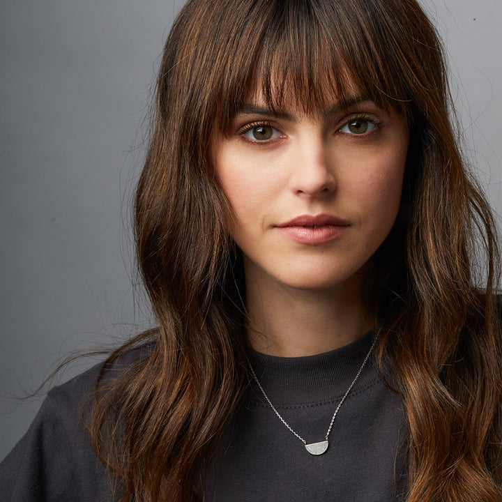 A woman with long brown hair and bangs is wearing a black shirt and the elegant Refined Neck Half Moon/Gold necklace from Scout Jewelry. The adjustable chain adds a touch of versatility, while the gray background provides a neutral, professional appearance.