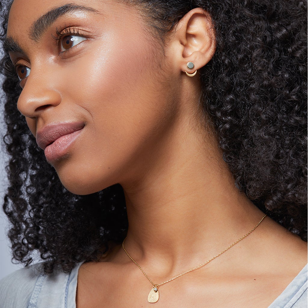 A woman with curly hair and a calm expression wears Scout Jewelry's Moon Phase Pyrite/Gold hypoallergenic earrings and a gold necklace with a semi-precious stone pendant, posing against a neutral background.