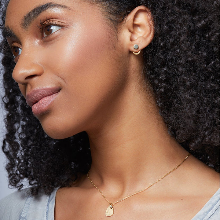A woman with curly hair and a calm expression wears Scout Jewelry's Moon Phase Pyrite/Gold hypoallergenic earrings and a gold necklace with a semi-precious stone pendant, posing against a neutral background.