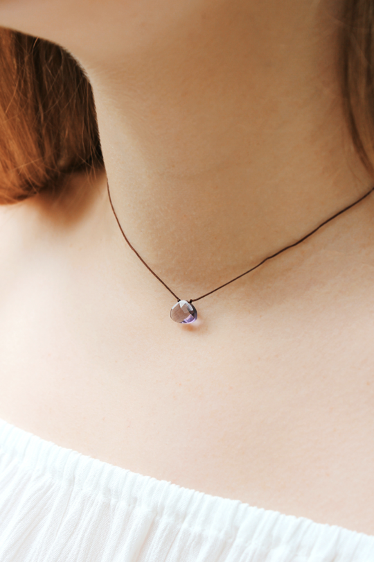 A close-up showcases a woman's neck wearing the "Power Necklace Violet Creativity" by LuckyFeather—a thin black cord necklace with a small violet teardrop stone pendant. Wearing an off-white, off-the-shoulder top against a softly blurred background, she exudes elegance and creativity.