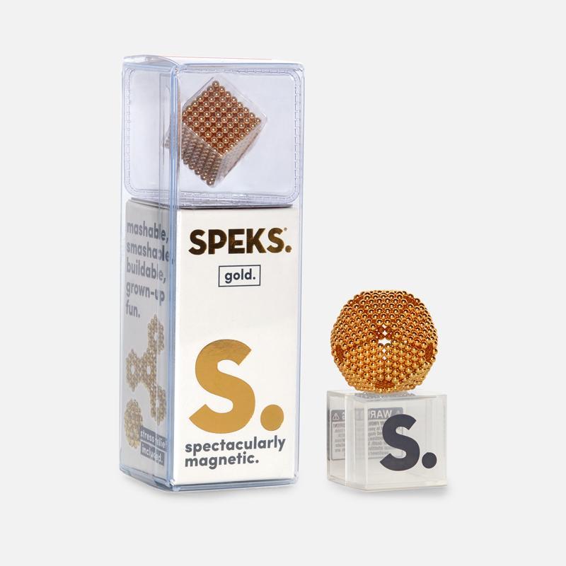 The image showcases the 512 Gold Speks, a set of gold-colored magnetic balls by SPEKS, ideal as a desk toy. The packaging highlights phrases such as "mashable, smashable, buildable, grown-up fun," and "spectacularly magnetic." One display ball is formed into a cube while the other is shaped like a sphere.