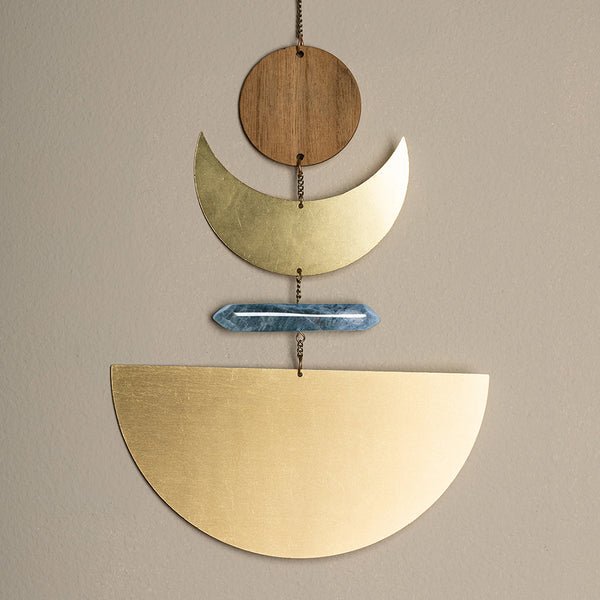 Introducing the Moonrise Labradorite Wall Hanging by Scout Jewelry: a stunning piece of sculptural wall art composed of geometric shapes, including a small wooden circle, two natural brass crescents, a blue crystal rod, and a large semicircle at the bottom, all elegantly linked by a delicate chain.