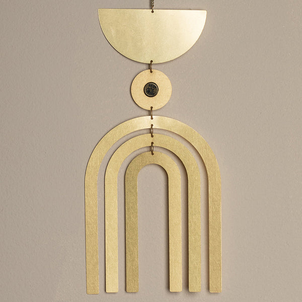 Introducing the Supernova Pyrite Wall Hanging by Scout Jewelry: a stunning piece of sculptural wall art showcasing a gold geometric design. It features a half-circle at the top, a small circle beneath it, and three arches gracefully descending against a simple beige backdrop.