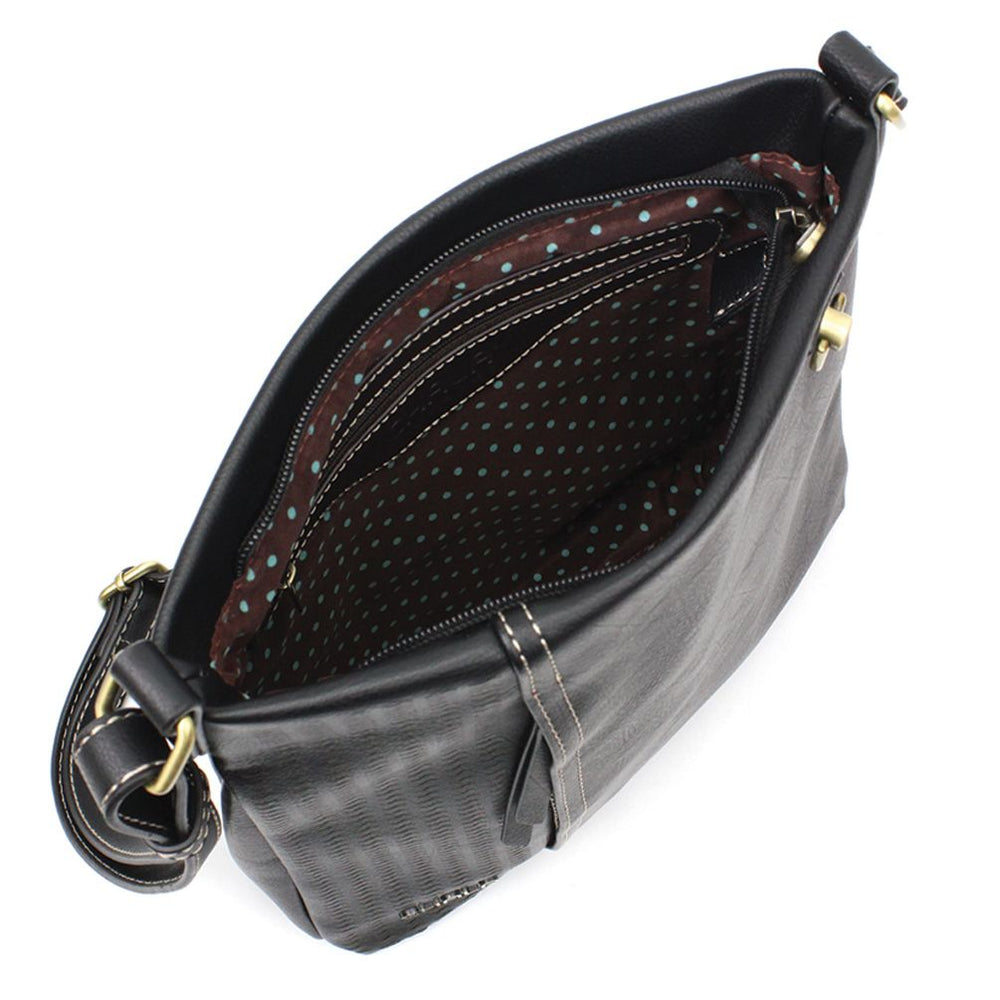 The Lasercut Crossbody DF Black by Chala is displayed from a top view, showcasing its black faux leather design with a polka dot fabric lining. The open bag reveals an interior zipper pocket and a main compartment. Its adjustable shoulder strap, embellished with brass buckles on the sides, enhances its versatile charm.