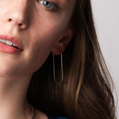 A close-up of a young woman with light skin and blue eyes, wearing a single long, thin, rectangular earring called "Cosmic Oval Silver" made of 925 sterling silver from Scout Jewelry on her left ear. She has long, brown hair and is looking slightly off to the side. The background is plain and white.