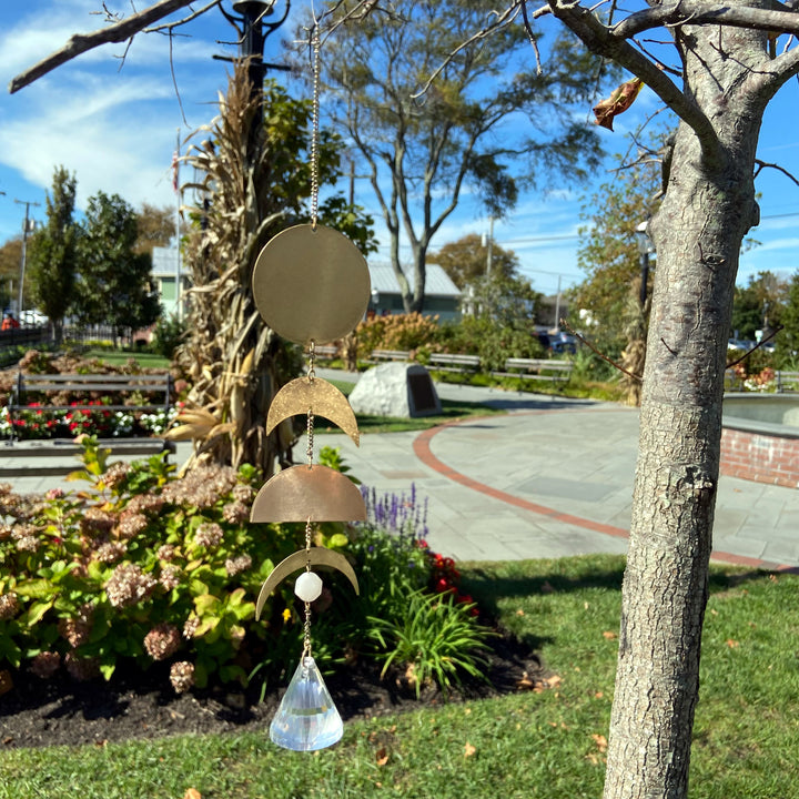 A Moon Phase Moonstone Suncatcher from Scout Jewelry is suspended from a tree branch in a park, its shimmering elements capturing the clear blue sky. Behind it, vibrant flower beds add color to the view, while a stone walkway and benches offer a place for relaxation. Delicate rainbows dance through the suncatcher's sparkling gemstones.