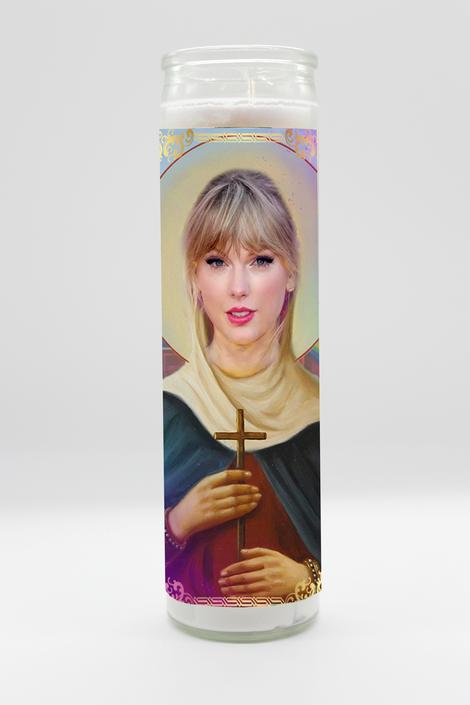 The Taylor Swift Candle by BOBBYK BOUTIQUE is a tall cylindrical, unscented candle with white wax that features an artistic depiction of Taylor Swift holding a cross. She has light-colored hair with bangs and is dressed in traditional saint-like attire with a halo behind her head, echoing the reverence of a celebrity shrine. The background is plain white.